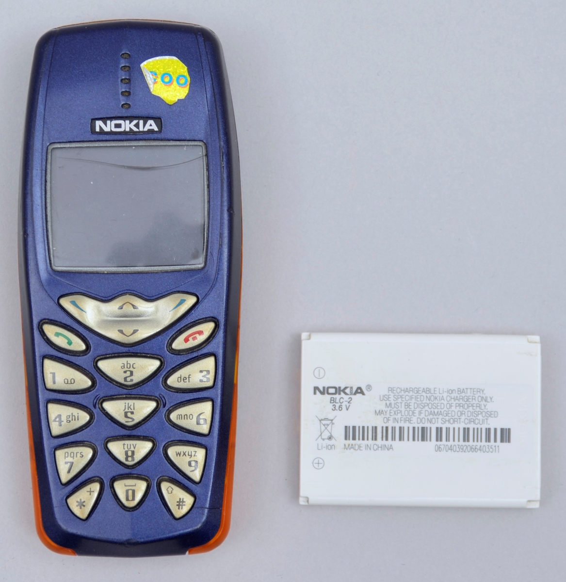 Rektangulær. Tastar og liten skjerm på eine sida. Blå med detaljar i oransj på sidane. Batteri.