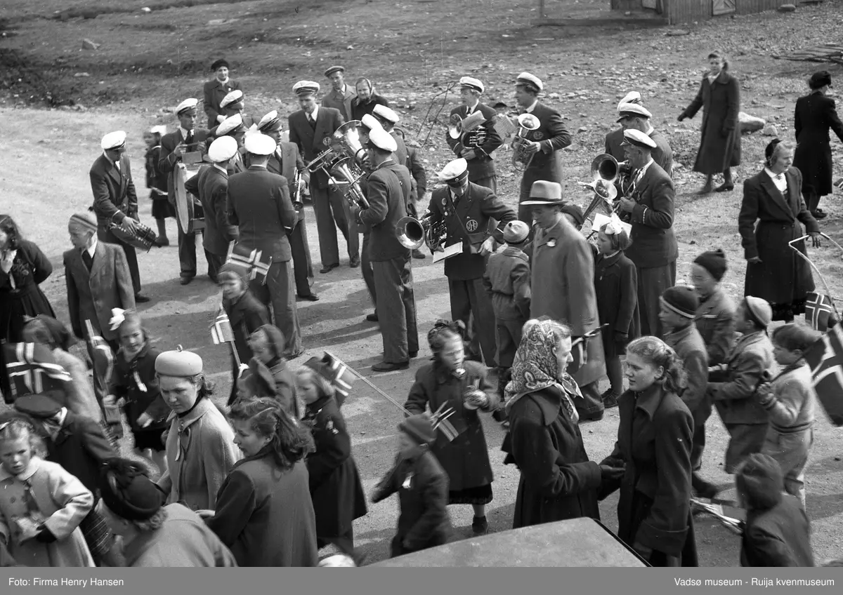 Det er pause i 17. maitoget i Vadsø 1951, vi ser barn, kvinner, menn og musikkorpsmedlemmer som står og snakker sammen.