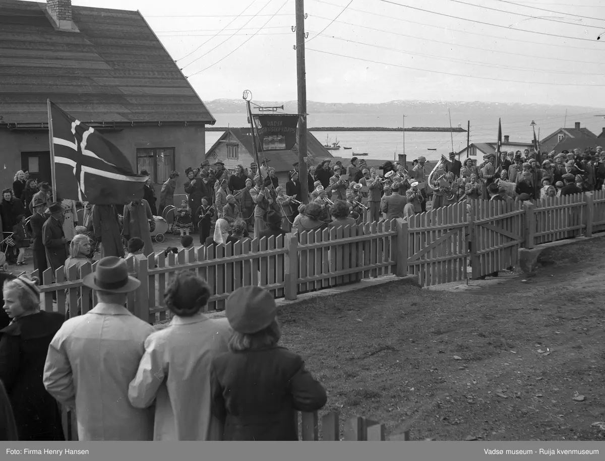 Vadsø sentrum 17. mai 1951. Nyborgveien, foran tidligere "Alders Hvile". Musikkkorps står oppstilt og spiller. (Fane: Vadsø Musikk-Korps). I tillegg ser man kvinnelige korsangere med sangerluer. Voksne og barn er tilskuere. Korpset står foran antatt baker Alf Hildonens bolig- og forretningsbygg. Vi ser flere bolighus og moloen i bakgrunnen.