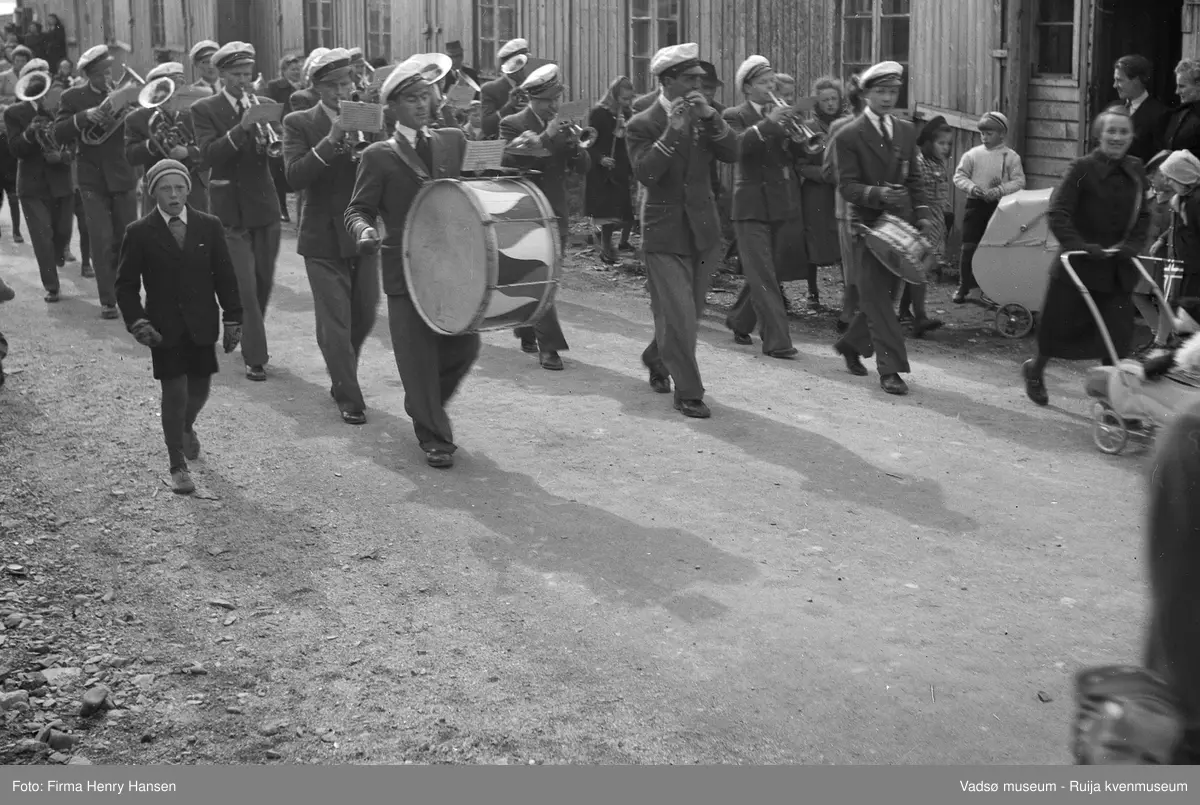 Vadsø sentrum 17. mai 1951. Musikkorps spiller i gatene. Trommeslagerne går fremst. Musikerne har uniformer og hvite hatter. Det står tilskuere ved husveggene og to barnevogner vises.