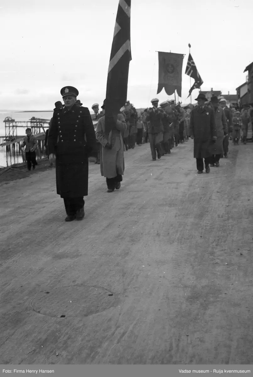 Vadsø 17. mai 1951. Musikkkorps masjerer i Indrebyen, antas å være i Havnegata. Vi ser flagg og fane. Fremst i toget masjerer en politimann som antas å være senere politimester Wickstrøm. Publikum, barn og voksne sees langs 17.maitoget. I venstre billedkant skimter vi sjøen med kaier, hjellbruk og nothjul.