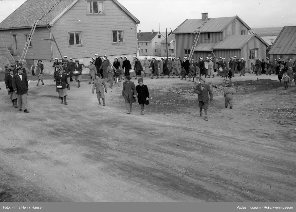 Vadsø 17. mai 1951. Musikkkorps og kor marsjerer opp Kirkegata og svinger mot vest innover i Amtmannsgata. Huset i venstre bakkant tilhørte Reidar Olsen, senere tjenestebolig for NRK-ansatte. Publikum, både barn og voksne ses langs 17.maitoget. Bygningen nede i Kirkegata i høyre billedkant er Bettenhuset og vi skimter også deler av havnen og Vadsøya.