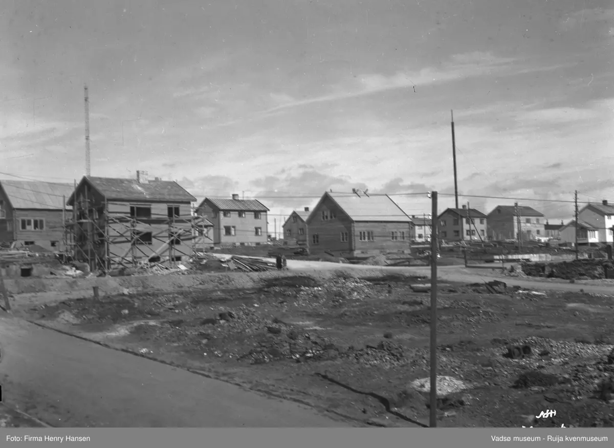 Bildet viser et kvartal med gjenreisningshus i Vadsø 1948. Øverst til venstre sees en av radiomastene. I forgrunnen sees en tomt med rester etter nedbrente hus, her ble hotellet oppført senere. Helt i venstre kant ser en kjellermuren av et nedbrent hus. Huset til høyre for ruinen er under oppføring og har stilas.  Eieren antas å være Olaf Hollum. Midt i bildet ser en Bettengården. Mellom Hollumgården og Bettengården krysses Kirkegata og Oscarsgate, den siste går på nedsiden av husene. Til høyre i bildet ser en branntomter på begge sider av Oscarsgate.