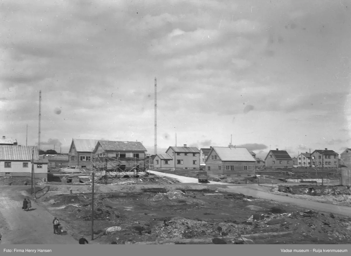Bildet viser gjenreisningshus i Vadsø sentrum 1948.  Tre radiomaster sees. Branntomta i forgrunnen antas å være hvor hotellet nå ligger.  Bakken sentralt i bildet antas å være Kirkegata der den krysser Oscarsgate.  Huset med stilas antas være Hollumgården (Olaf Hollum), det laftede huset på andre siden av bakken er Gjertrud Betten eide i seinere år. I venstre hjørne sees to personer som leier sykkelen i bakken og to kvinner som går. Denne bakken er borte etter byregulering.