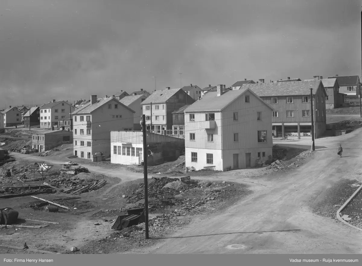 Vadsø sentrum 1950-tallet. Fotografert mot nordvest. Veikrysset er mellom Tollbugata nordover, Kaigata og Havnegata mot vest. Ned mot Kaigata med inngang fra Havnegata ligger (sett østfra): Saxi, Helfjords trykkeri, bokhandelen og Georg Evangers stall (senere Gundersens fiskebutikk). På nordsiden av Havnegata ligger (sett østfra): Ekviperingen, et bygg med høy sokkeletasje. Ved siden av hadde Rolf og Henry Hansen butikk, i boligdelen bodde Rolf Hansens familie.