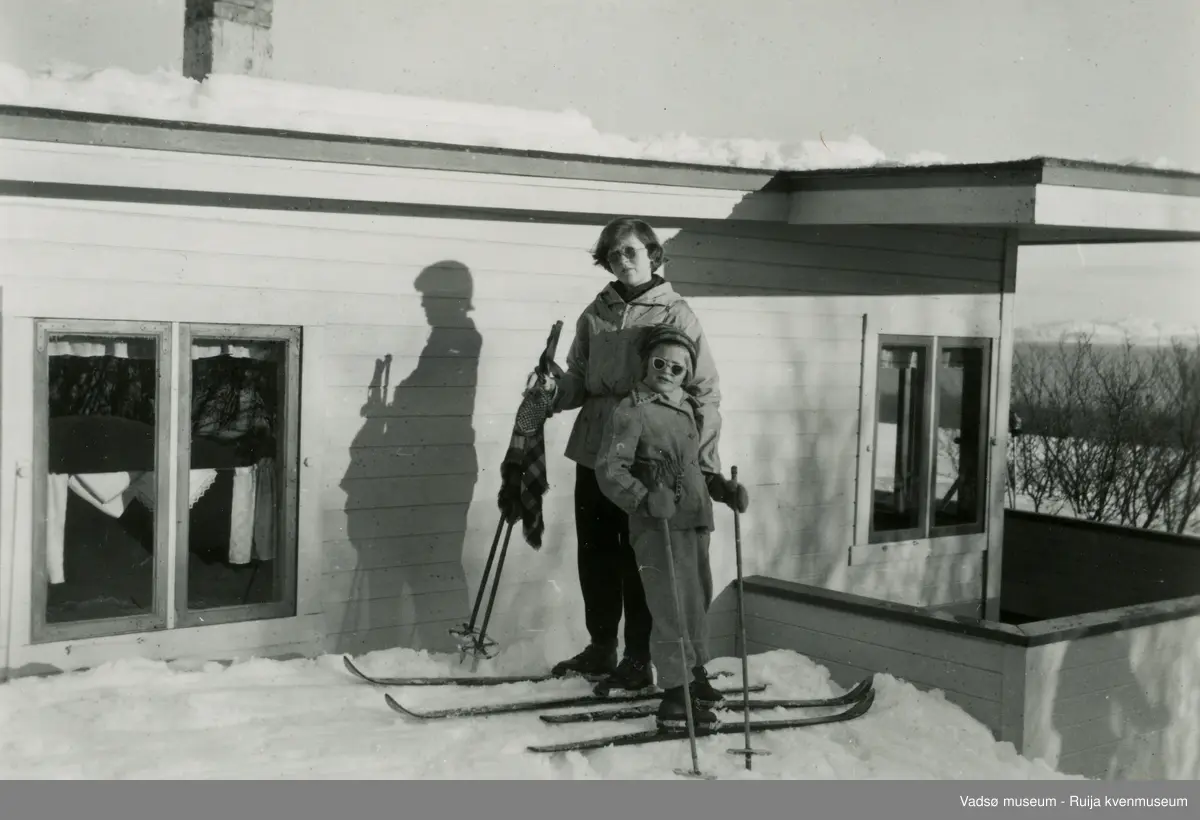 Fra venstre ser vi Liv og Åse Hansen (søstre) utenfor deres familiehytte i Laksebybukt utenfor Vadsø på 1950-tallet. Varangerfjorden i bakgrunnen.