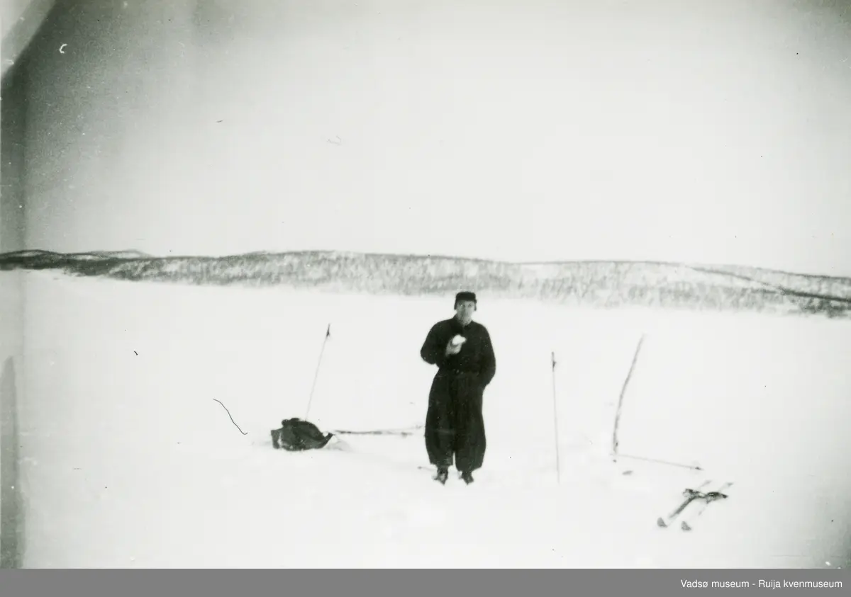 Mann sannsynligvis på isfiske, inntar sin nistemat. Vi ser kjelke/slede, ski og skistaver. Åsside med litt skog i bakgrunnen.