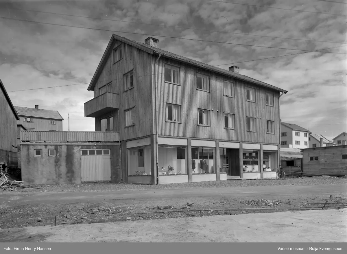 Havnegata 1 i Vadsø med gjenreisingsbebyggelse. Bygningen er Ekviperingen, med forretning i sokkeletasjen og boliger i 1. og 2.etasje. Huset har skifertak.   I venstre kant av bygget, mot vest er det en takterrasse over en garasje i sokkelen. I andre etasje en luftebalkong. Bak takterassen sees en radiomast og deler av en bygning i Nyborgveien. I forretningslokalet, I utstillingsvinduene i sokkeletasjen ser man herrekonfeksjon og sko. Foran bygget er det et sykkelstativ med en parkert sykkel. I høyre billedkant ser vi en brakke og et skur i forgrunnen og Niskagården og to andre bolighus i Nyborgveien. I forkant av bildet er en grunnmur.
I dag huser første etasje advokat Hegg.