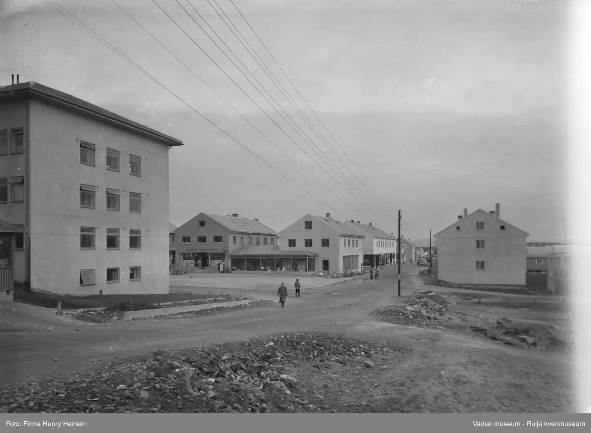 Vadsø Sentrum på 1950-tallet. Bildet er tatt fra nord nedover Tollbugata mot sør. Helt i venstre billedkant ser vi deler av Kiosken og vestveggen på Rådhuset. Foran Rådhuset ligger Torget hvor vi ser sykler, barnevogn og mennesker foran forretningsbyggene til Henry Hansen og Rolf Hansen. I Tollbugata ses folk som spaserer eller sykler. Det første bygget i Tollbugata er R. G. Esbensens butikk. Nederst i Tollbugata ser vi et par biler.  Til høyre i bildet ses noen hauger med jord og stein.