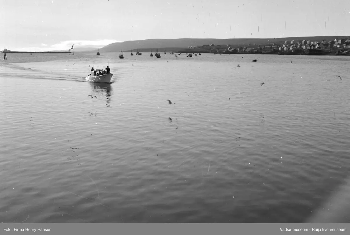 Kongebesøk i Vadsø i 1946. Bildet er tatt mot nordvest. Vi ser havna som ligger speilblank og deler av Indrebyen. Flere fiskebåter og småbåter ligger for anker. Sjaluppen med kongen og hans følge er på vei inn mot dampskipskaia. Skipet som kongebesøket kom med er ankret opp bak moloen.