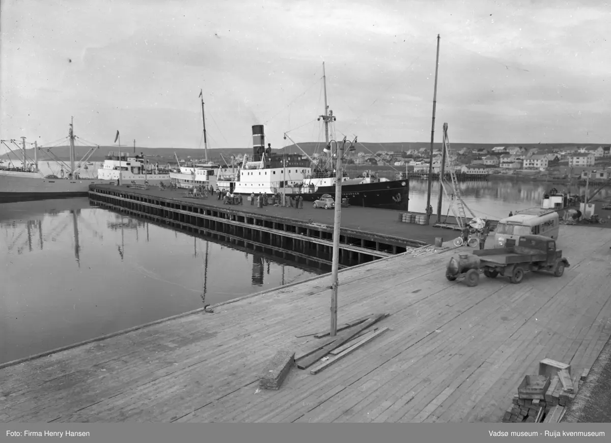 Vadsø 1952. Dampskipskaia. Bildet er tatt mot nordvest. Hurtigruteskipet "DS Finmarken" ligger ved kaia på nordsida. Nordenfjeldskes godsbåt "Guttorm Jarl" ligger ved kaia mot vest. På kaia nærmest kamera står en buss og en lastebil, lastebilen har tilhenger med en tønne på. Ved hurtigruta er det flere mennesker, og en personbil står parkert.