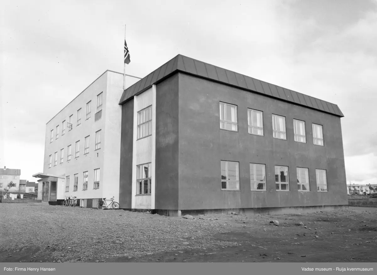 Vadsø 1957. Bildet viser Vadsø Samfunnshus og er tatt mot nordøst. Det står parkerte sykler langs veggen mot vest. I bakkant av bildet, på begge sider av samfunnshuset ser vi litt av bebyggelsen langs Idrettsveien. Flaggstangen på samfunnshuset er heist med flagget på halv stang.