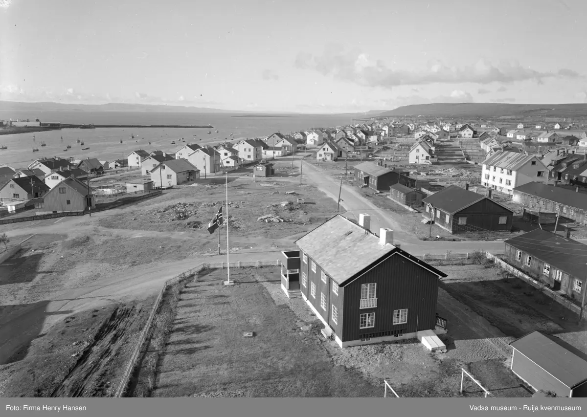 Vadsø 1957. Bilde tatt fra kirken mot vest. Prestegården nærmest. Det flagges på halv stang. Tverrgata er Fossevegen som ender i Damsveien. Veien mot vest finnes ikke lenger. I området foran ble Statens Hus senere bygget. Man ser brakker fra gjenreisingstida. Vågen med molo og fiskebåter. Vadsø Sildoljefabrikk (etablert i 1953) synes ved enden av moloen.