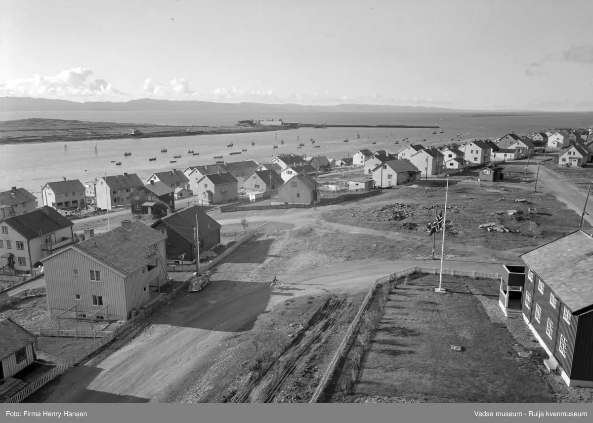 Vadsø 1957. Bilde tatt fra kirken mot vest. Prestegården nærmest. Det flagges på halv stang. Tverrgata er Fossevegen som ender i Damsveien. Det er en del brakker på bildet. Vågen sees med fiskebåter og molo. Vadsøya med Vadsø Sildoljefabrikk (etablert  i 1953).