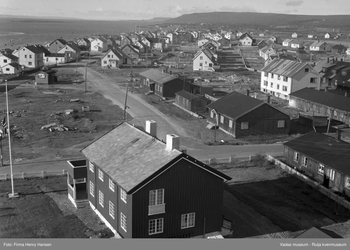 Vadsø 1957. Bildet er tatt fra kirken, om sto ferdig i 1958, mot vest. Vi ser prestegården i forkant av bildet med flagget på halv stang. Langs veien midt i bildet en del brakker fra gjenreisningstiden. Denne veien er sannsynligvis senere regulert bort. Til venstre i bildet bolighus langs Damsveien og fjorden med fiskebåter og Vadsøya i bakgrunnen. Til høyre og lengst fra kamera ses fiskehjeller rett bak bebyggelsen.