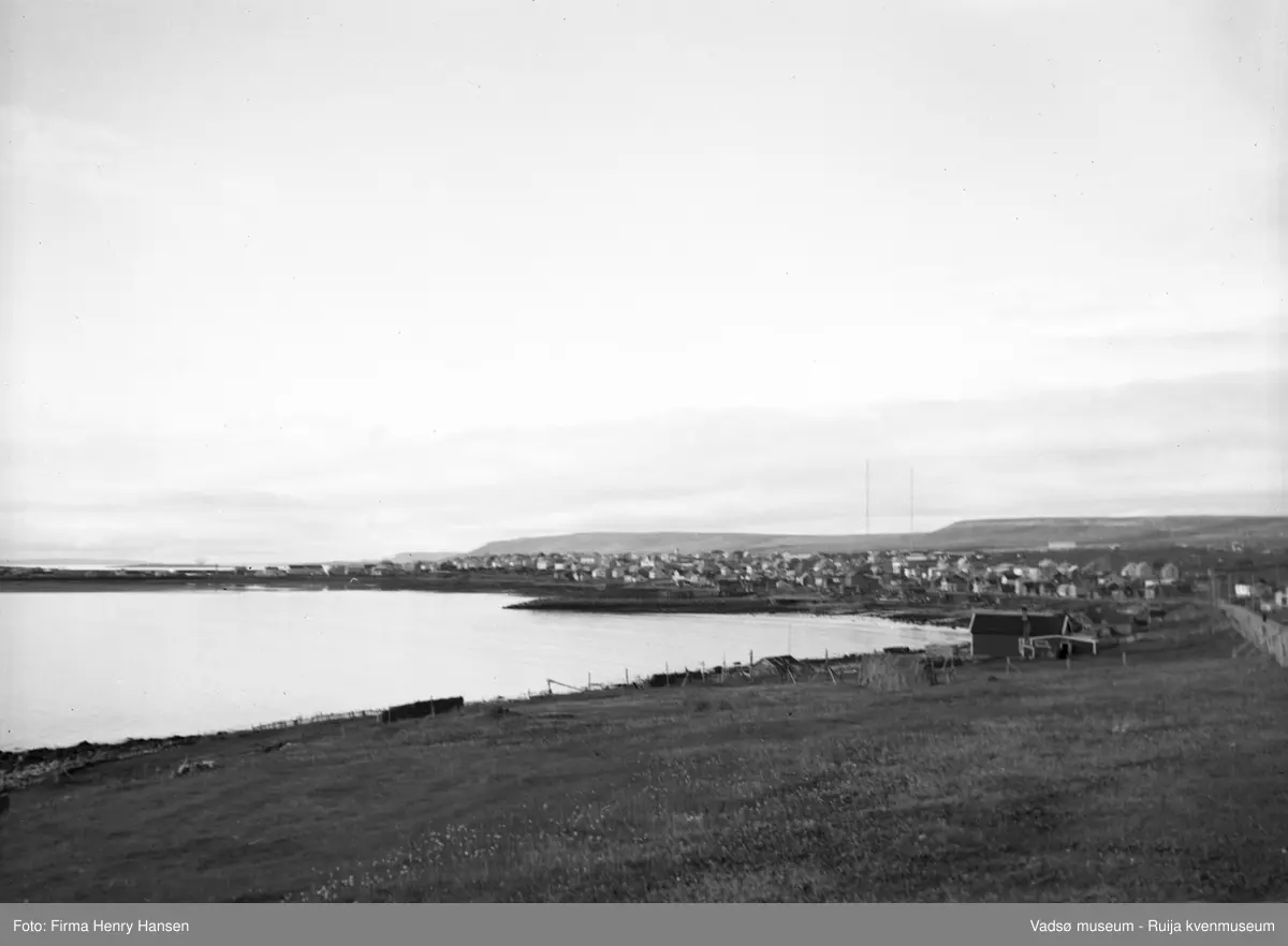Bildet viser Ytrebyen i Vadsø øst for Landhuselva sett fra øst 16.9.1951. I forgrunnen sees et jordstykke med hesjer. Vest for jordet er det inngjerdede bygninger.  Det framste antas å være et våningshus fordi det har skorstein. Det er muligens et varangerhus med lavere fjøs. Det er også flere mindre uthus på tomta. Bak disse bygningen ses et varangerhus. Huset bak dette igjen er også et varangerhus. På øversida av husene ser en Vardøveien. Videre ses flere eldre hus langs veien mot byen. Midt i bildet ses både eldre og nyoppførte hus. I bakgrunnen ses de to radiomastene. Til venstre i bildet ser en Ørtangen.