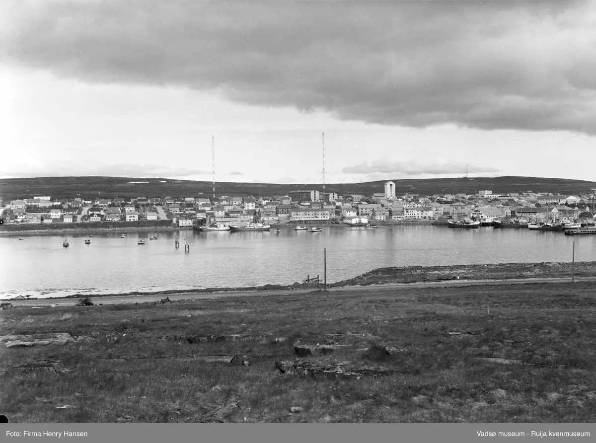 Motivet viser vestre del av Vadsø sentrum og havn sett fra Vadsøya 20.07.1968. Øverst i midten av bildet ser en de to radiomastene, Fylkesbygget, Statens hus, prestegården og Vadsø kirke. Andre offentlige bygg som synes er Vadsø rådhus og Vadsø skole. Helt til høyre i bildet sees ytterste del av dampskipskaia. På havna og ved kaianleggene sees store og små fiskebåter.  I forgrunnen ser man en del av veien som går fra brua til Sildoljefabrikken på Vadsøya.