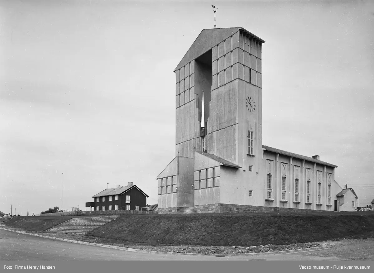 Vadsø kirke fotografert 27.07.1961. Bildet er tatt fra sydøst og vi ser syd- og østsiden av kirken. Foran kirken, Amtmannsgata med den brede trappa opp mot kirka. Til høyre for kirken kan vi se litt av Aug. Trastisgate. Til venstre for kirken ser vi Vadsø Prestegård med gjerde rundt eiendommen..
