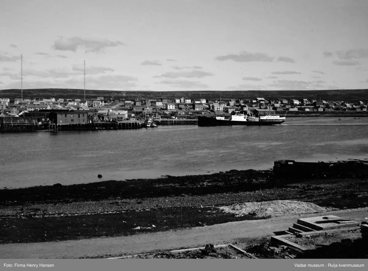 Vadsø havn. Hurtigruten D/S Finnmarken (eid av rederiet Vesteraalens Dampskipsselskap) legger til ved dampskipspiren. Indrebyen med bebyggelsen langs Havnegata og radiomastene bak. Bildet tatt fra Vadsøya, vest for broen.