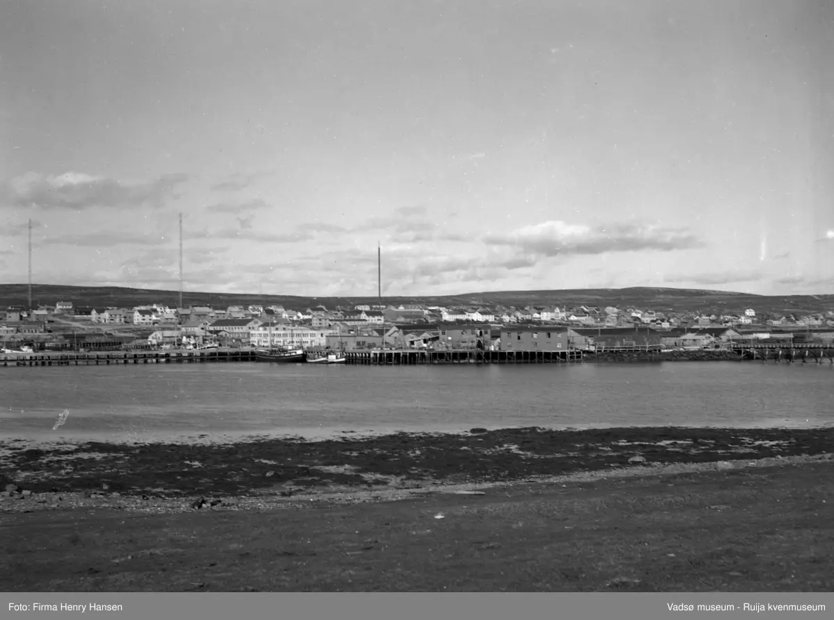 Oversiktbilde Vadsø Sentrum med  deler av Ytrebyen. Bildet er tatt fra Vadsøya mot nordøst. Vi ser havna med piren på dampskipskaia, dampskisekspedisjonen og et fiskebruk midt i bildet. Fiskebåter ligger fortøyd ved kaia. I høyre billedkant ser vi deler av brua over til Vadsøya. Videre ser vi Vadsø sentrum/Midtbyen og deler av Ytrebyen med gjenreisingsbebyggelse. Til venstre i bildet ser vi de to radiomastene. Midt i bildet en ukjent mast.