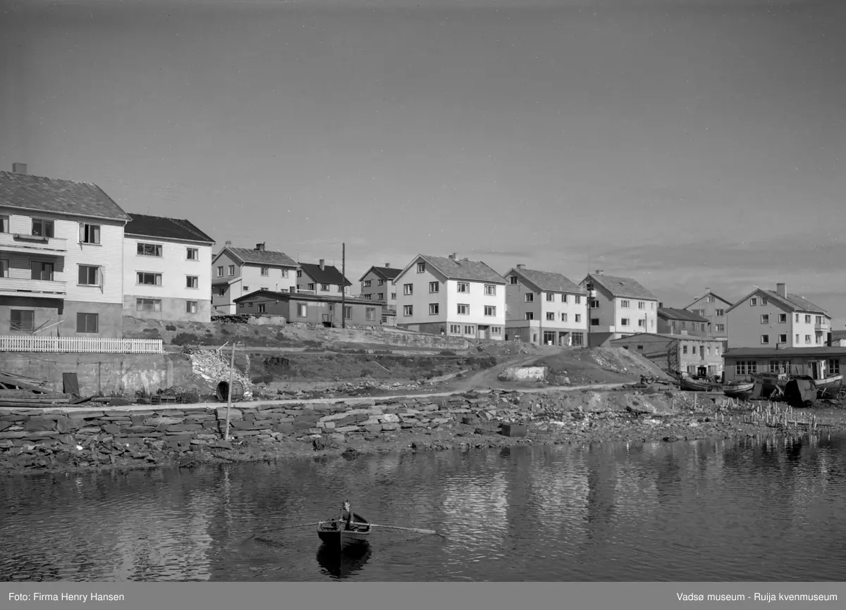 Oversiktsbilde over midtbyen i Vadsø og deler av Indrebyen med en speilblank havn i forgrunnen. I forgrunnen ser vi en ung gutt i robåt. Det er en tørrmur fra fjæra og opp mot nederste gate. Ruiner, rester etter bygninger og kaier ødelagt under 2.verdenskrig. I høyre billedkant brakker og fiskebåter. Videre ser vi ser flere gjenreisingsboliger med næringsvirksomhet i kjelleretasjen.