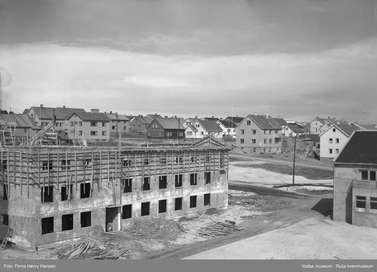 Vadsø 3.6.1951, Sentrum. Bildet er tatt fra sørvest, motivet er først og fremst Rådhuset under bygging. Fasaden mot sør er påbegynt, første og andre etasje er ferdig murt med åpninger for vinduer, i tredje etasje er forskalingen på. Bildet viser en del av torget, og en del av baker Henriksens bygg vises til høyre i bildet.