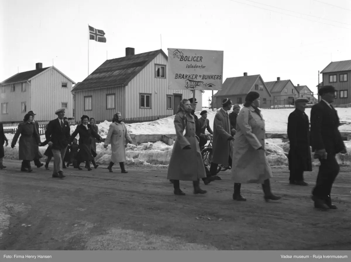 Vadsø 1.mai 1952, Oscarsgata. Huset midt i bildet er Natalie Øyens bolig. Folk, kvinner, barn og menn, går i tog. En mann bærer en plakat med tekst "Boliger istedenfor brakker og bunkers" . På plakaten er det også to tegninger av hus. I bakgrunnen ser vi flere bolighus, flaggstang med norsk flagg ved ett av husene. Det er snø på bakken.