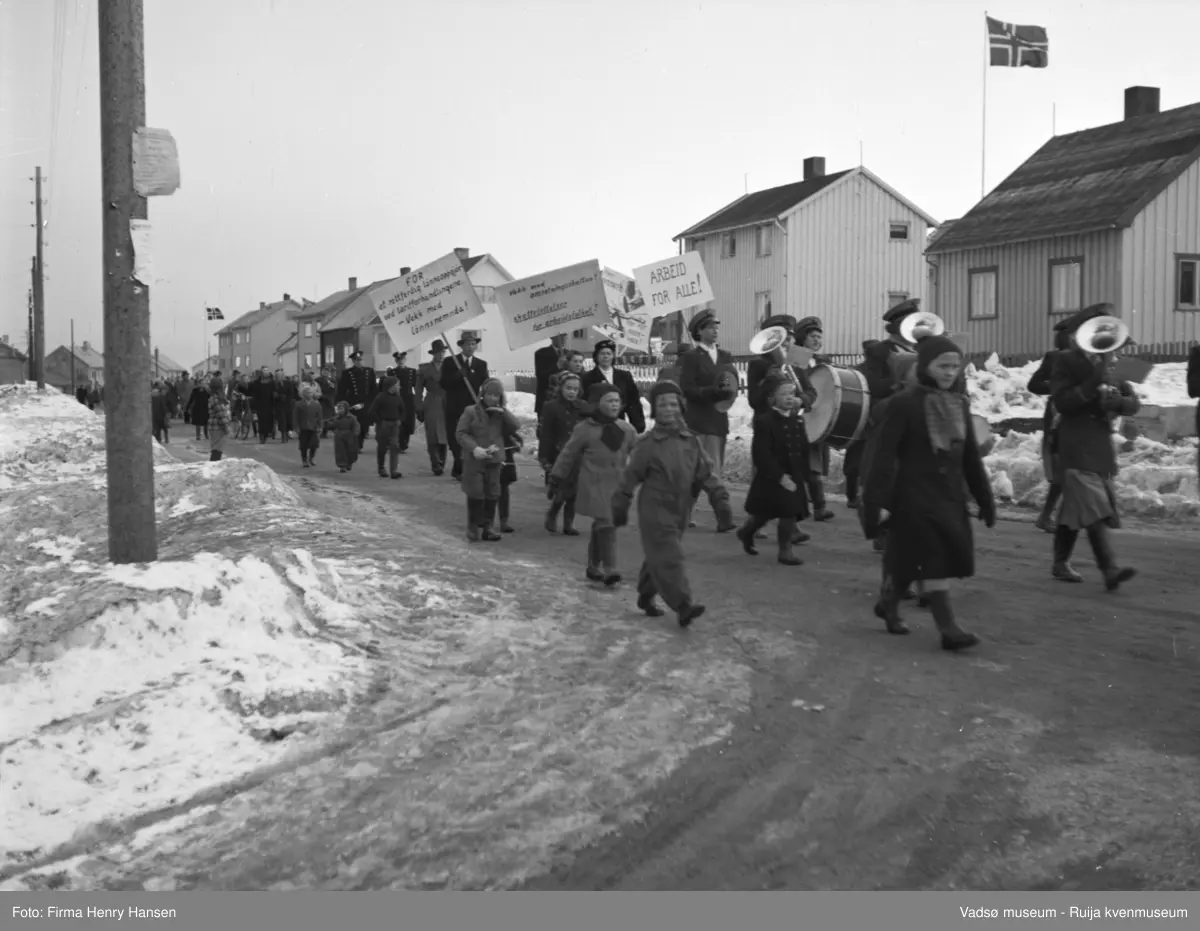Vadsø 1.mai 1952, ved krysset Oscarsgata-Kirkegata. Mange mennesker - kvinner, menn, barn - går i demonstrasjonstoget. Noen bærer musikkinstrumenter. I bakgrunnen flere bolighus, flaggstang med norsk flagg ved to av husene. To politimenn i uniform i toget. Tre menn bærer hver sin plakat, med disse tekstene: "Arbeid for alle",  "For et rettferdig lønnsoppgjør", "Vekk med omsetningsskatten". Snø på bakken.