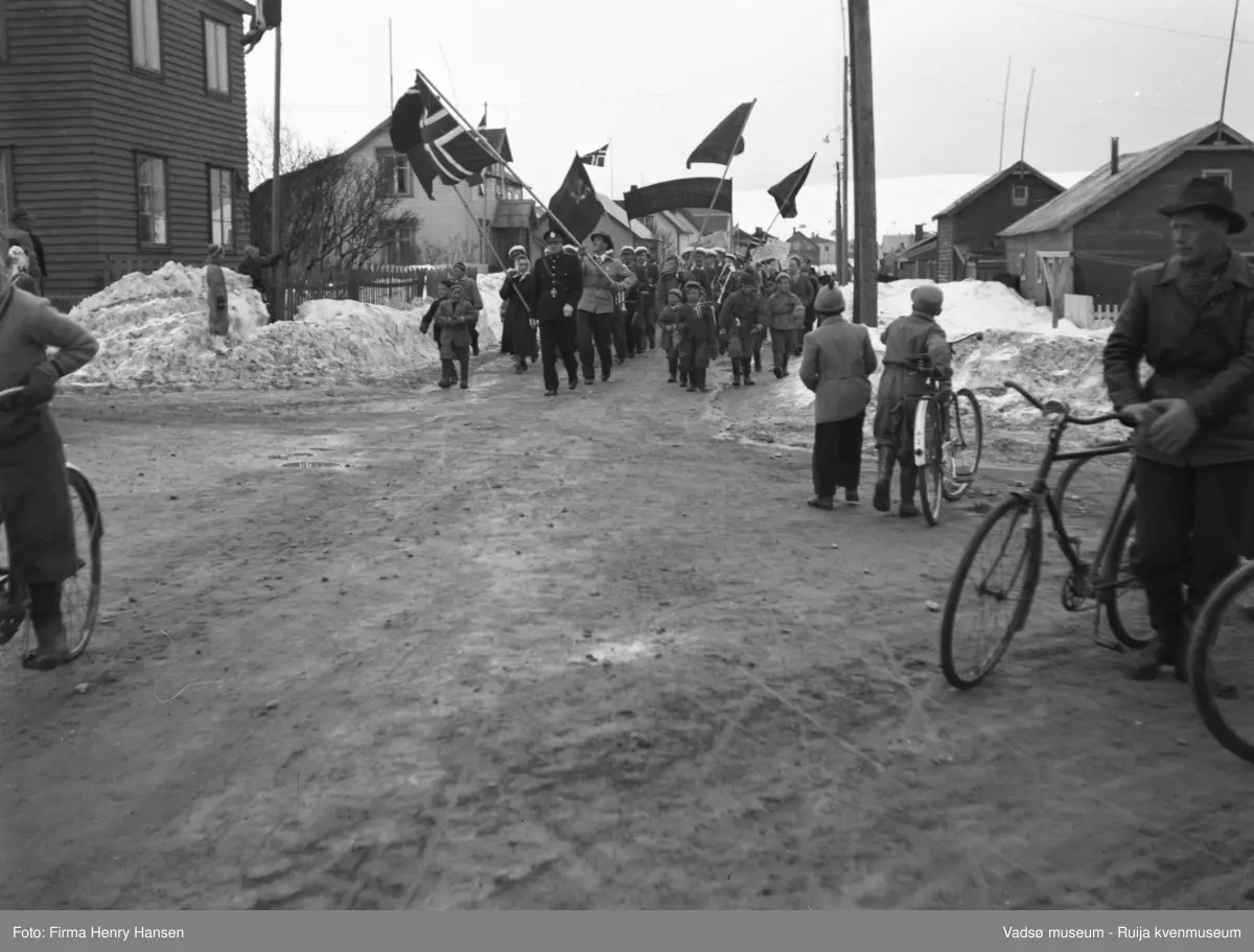 Vadsø 1.mai 1952. Bildet viser 1.mai-toget. En politimann i uniform,  går først i toget, bak ham kvinner og menn, mange menn med hvit lue/ caps på hodet. Flere flagg, bl.a. et stort norsk flagg og et flagg med hammer og sigd. I forgrunnen en mann med sykkel, og to gutter med en sykkel.  Det er snø på bakken.