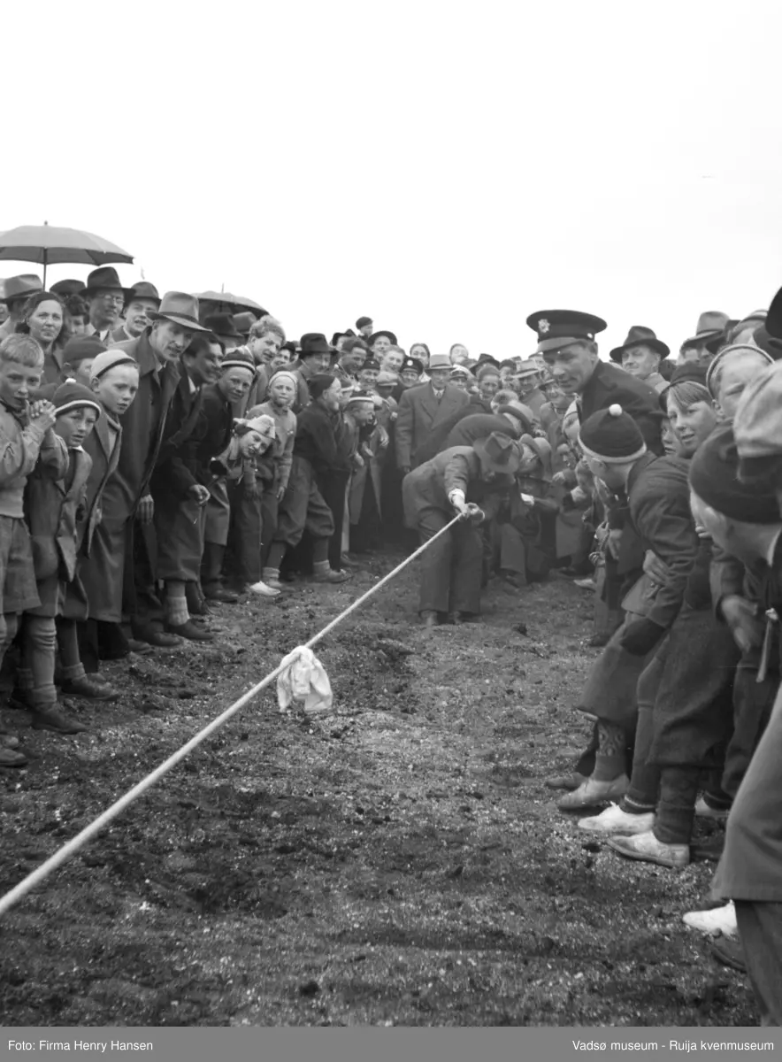 Vadsø 17 mai 1951. Tautrekkingskonkurranse. I enden av tauet ser vi menn i frakk og hatt som trekker i tauet. En stor menneskemengde, bl.a. en politimann i uniform, står på begge sider av tauet og følger med konkurransen.  I menneskemengden ser vi bl.a. menn med hatt og frakk, speidere, gutter i toppluer, paraplyer, og folk med uniformslue, studenterlue og sangerluer.