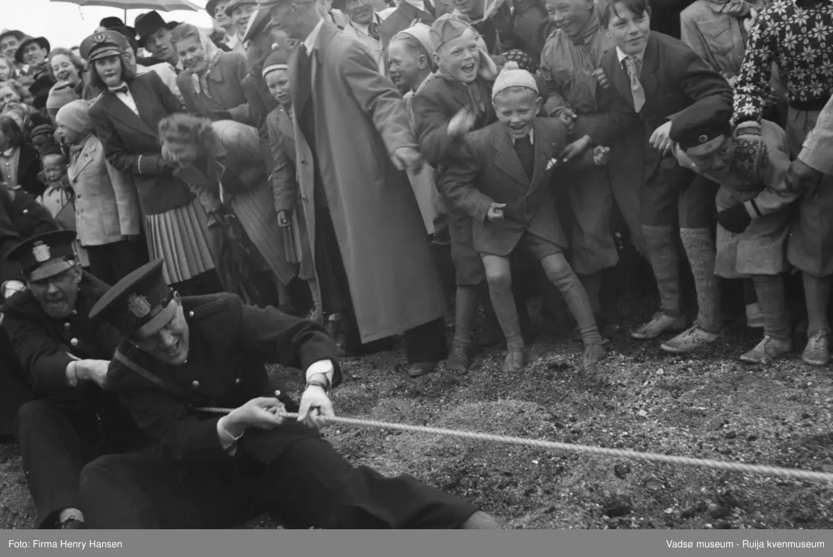 Vadsø 17 mai 1951. Tautrekkingkonkurranse. I enden av tauet ser vi politimenn i uniform som trekker i tauet. En stor menneskemengde står og bivåner det hele. I  menneskemengden ser vi bl.a. menn i hatt og frakk, speidere, jente i korpsuniform, damer med tørkle, paraplyer og gutter med toppluer.