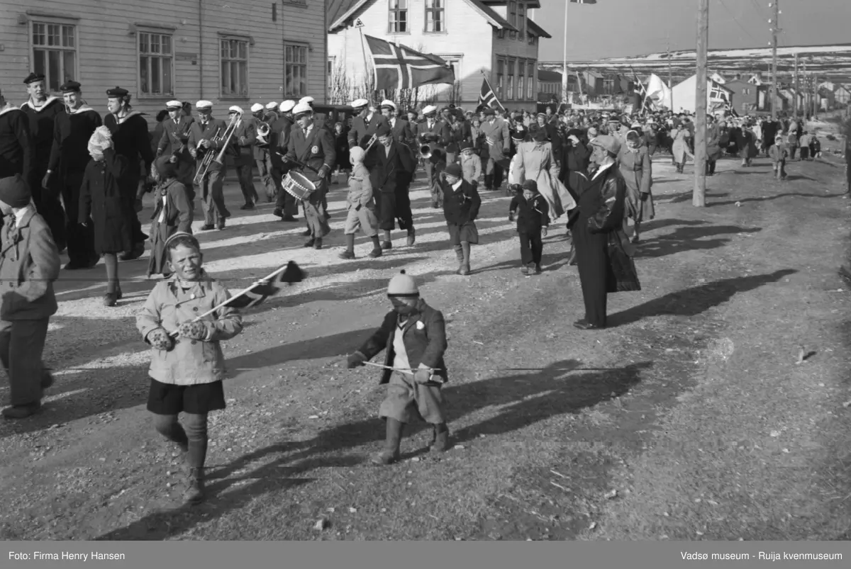 Vadsø 17 mai 1951. Bildet er tatt mot øst. 17.maitoget, muligens folketoget, marsjerer i Ytrebyen. I høyre bakkant av bildet ser vi Melkevarden, litt snedekket,  I venstre billedkant, foran i toget, ser vi noen marinegaster. Rett bak kommer musikkorpset marsjerende. Folk og barn med flagg sees i og langs toget,