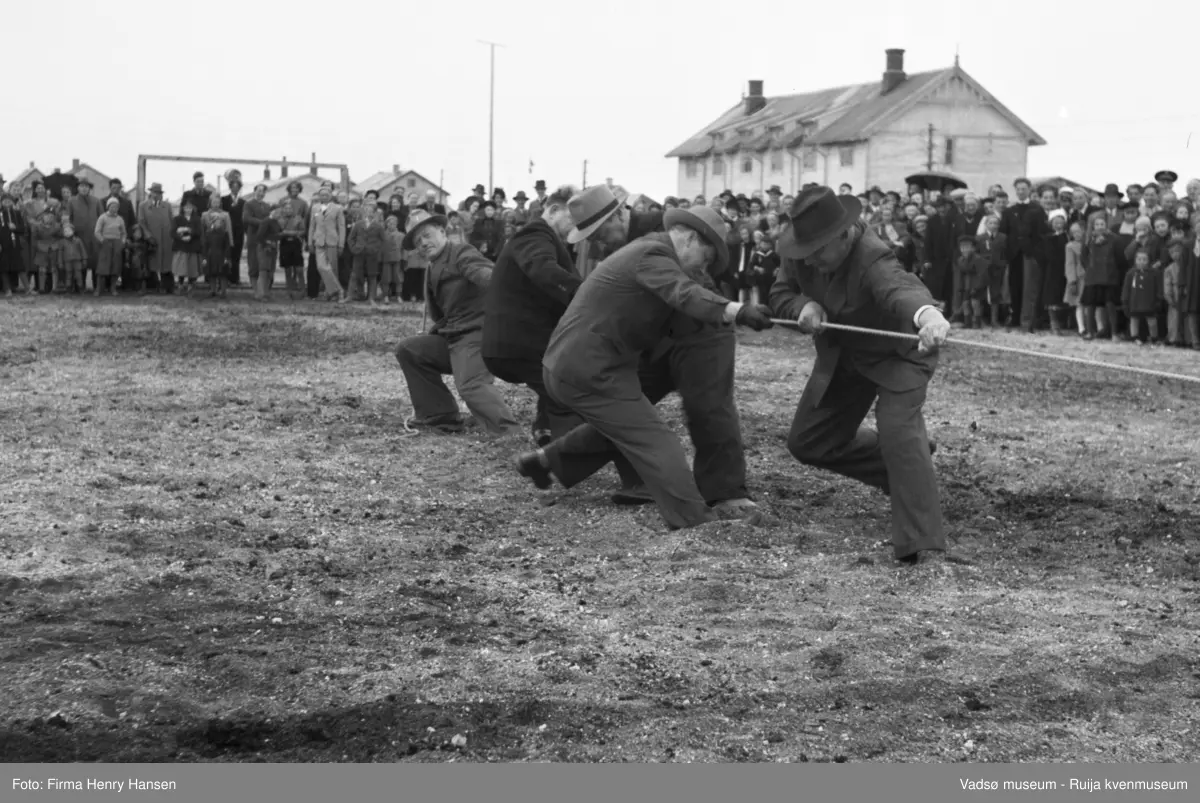 Vadsø 17 mai 1951. Tautrekkingkonkurranse. I enden av tauet ser vi fem menn, fire med hatt, som trekker i tauet. En stor menneskemengde, barn og voksne, kvinner og menn, står og bivåner det hele.  I bakkant av bildet ser vi deler av en større bygning, annen etasje og taket med to piper samt flere bolighus.