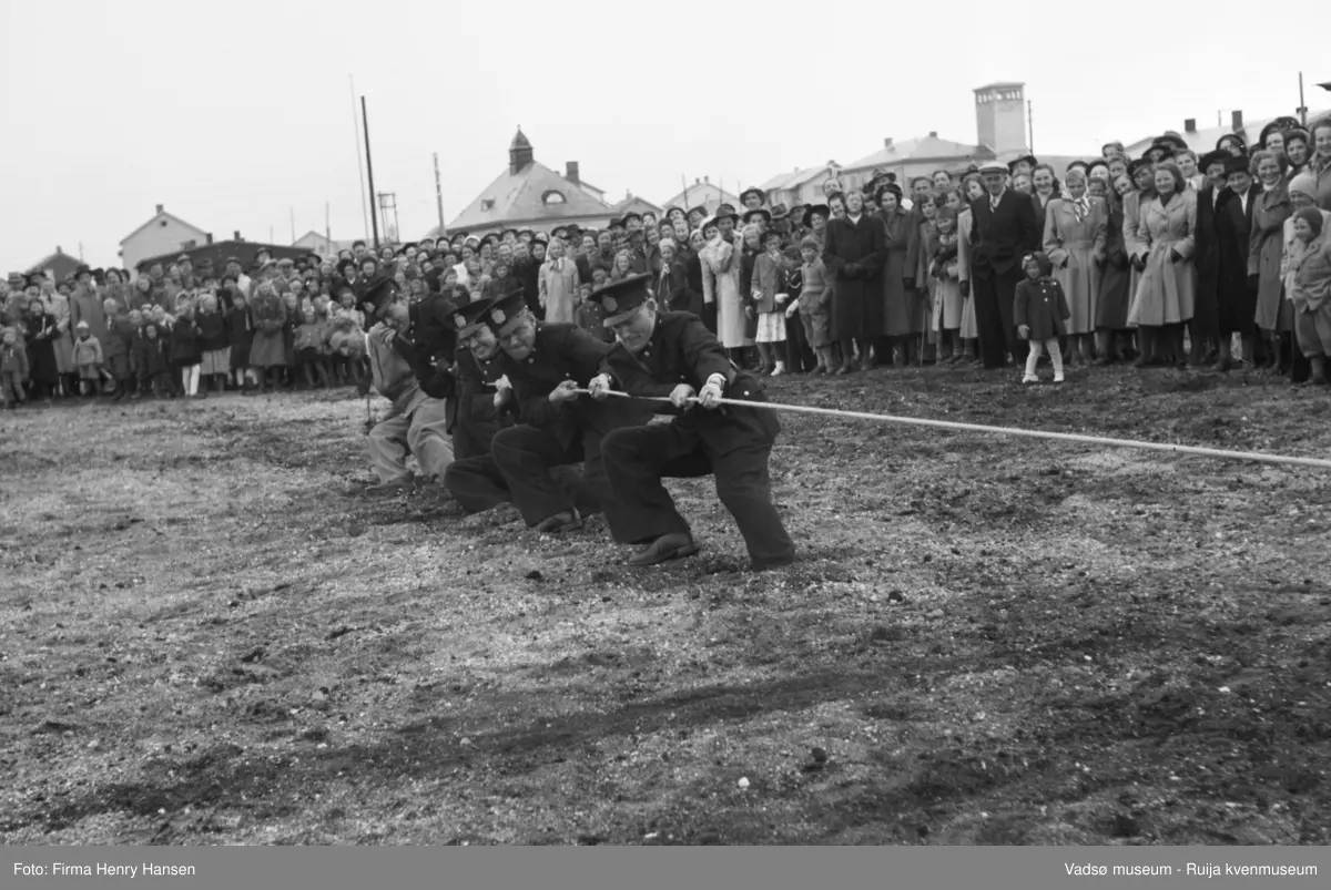 Vadsø 17 mai 1951. Tautrekkingkonkurranse, muligens på idrettsplassen. I enden av tauet ser vi fem menn, fire i politiuniform, som trekker i tauet. En stor menneskemengde, barn og voksne, kvinner og menn, står og bivåner det hele.  I bakkant av bildet ser vi bygninger, bl.a. tårnet og deler av brannstasjonen og taket til Finnmarksbiblioteket.