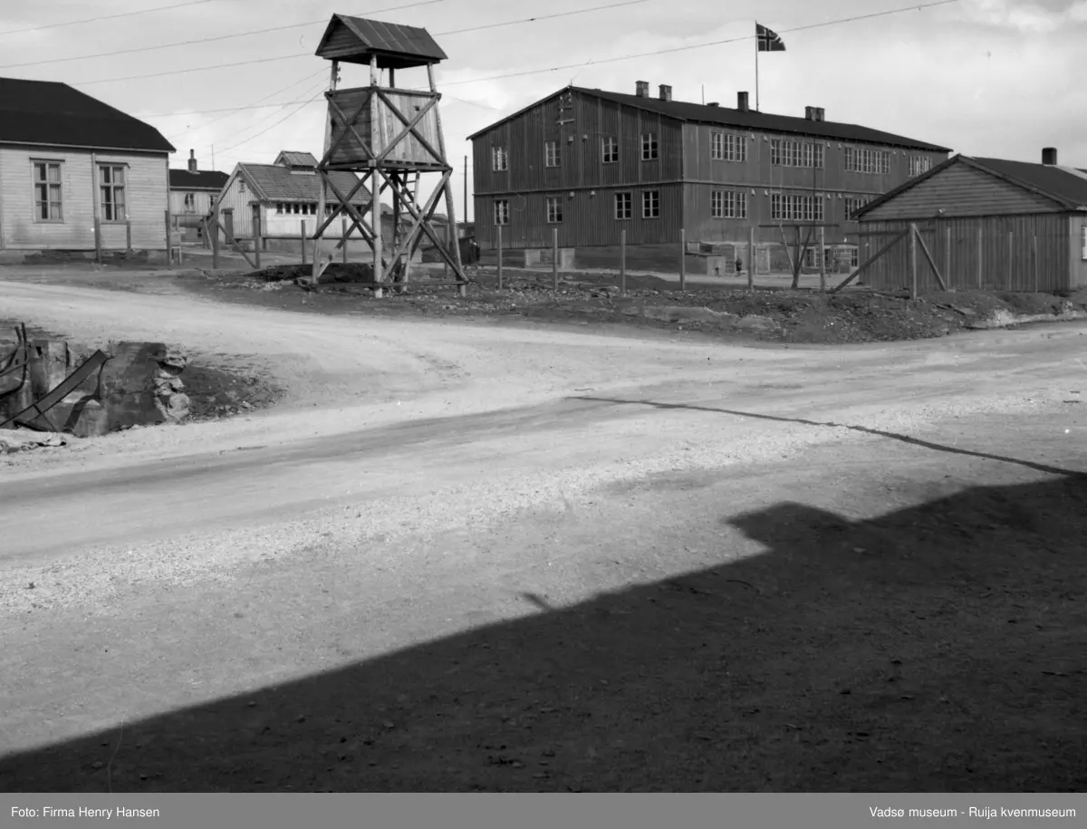 Vadsø sentrum. Bildet er tatt mot nordøst. Helt i forgrunnen ser vi krysset Oscarsgate - Skolegata. Vi ser en stor toetasjes bygning med stor grunnmur, mange vinduer i begge etasjer og i grunnmuren. Over taket er et norsk flagg heist. Bygningen er Brakkeskolen, en provisorisk skolebygg reist av Staten v/ Finnmarkskontoret,  Sør for skolen vises litt av en eneetasjes bygning. I forgrunnen, vest for skolen står det et tårn av tre, antagelig et klokketårn. Bak tårnet del av en bygning, det er "Det gamle bedehus" for den læstadianske menighet. Fra 2.verdenskrig til 1958 ble huset brukt som menighetskirke, derfor klokketårnet. I bakgrunnen ser man to andre bygninger, det ser ut som eldre bygninger, ikke gjenreisningshus.