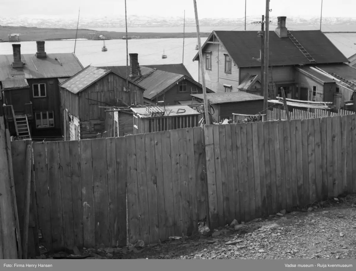 Vadsø, Indrebyen. Bildet er tatt mot sør, i bakgrunnen ser man litt av havna og vestre del av Vadsøya med moloen i vest. Man ser også litt av Varangerfjorden og land på sørsida av fjorden. Helt i forgrunnen på bildet er det et høyt plankegjerde. Bak gjerdet flere bygninger, de har fasaden mot Havnegata, fotografen har tatt bilde av husene inn mot bakgården. Man ser hustakene og bare litt av resten av bygningene. Tre av bygningene er trolig bolighus, flere mindre uthus. Alle byggene er gamle, fra før krigen, de står tett ved hverandre. På havna liggere tre fiskebåter, på øya ser man to kaier, en bygning ved hver kai, og en båt som ligger i fjæra lenger mot vest.