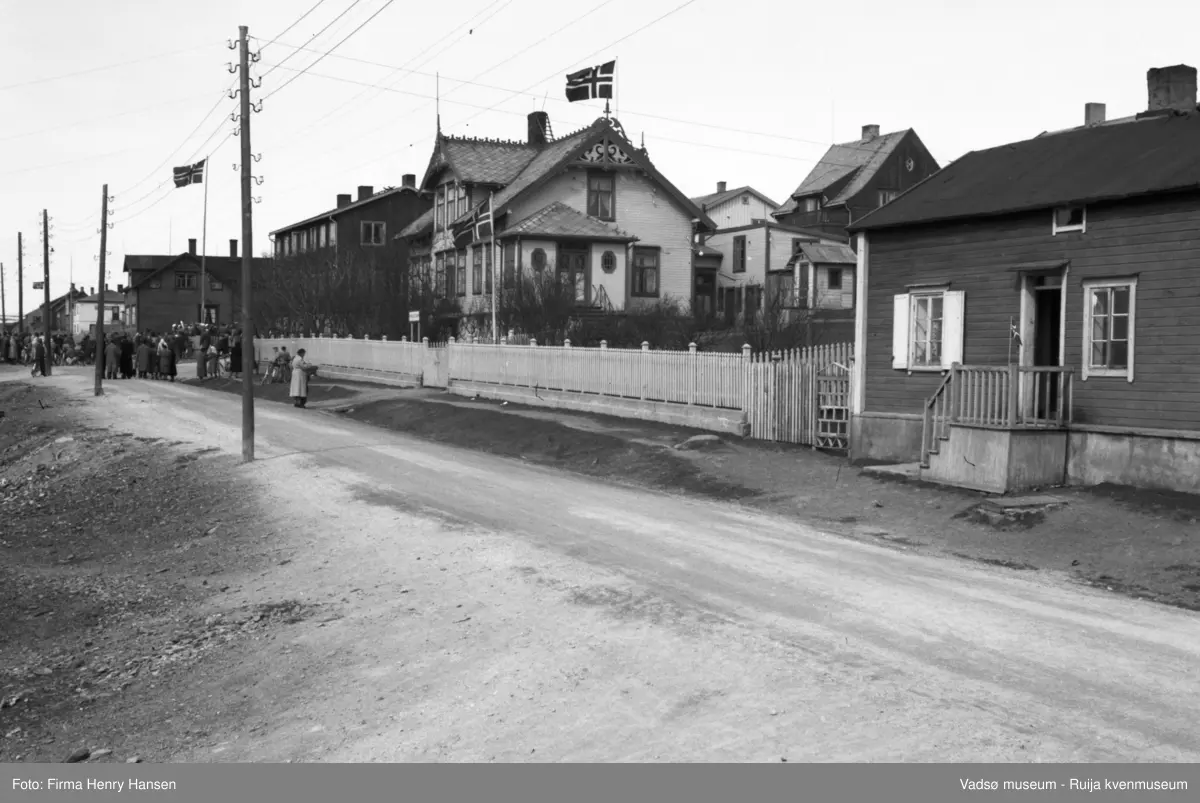 Vadsø, Nyborgveien. Bildet er tatt mot nordvest, med gataen i forgrunnen. Det er flere bygninger langs gaten og flagg ved tre av husene. Folkemengde i bakgrunnen, i krysset ved gamlehjemmet Alders hvile, kanskje det er 17.mai. Midt på bildet er Esbensens hus "på Bakken", Nyborgv. 33, med hvitmalt stakittgjerde. Flaggstang med flagg både ved Alders hvile og Esbensens hus. Et lite enetasjes hus helt i forgrunnen, med trapp ut mot gata. Bak Esbensens hus ser man litt av to bygninger i Damsveien, blant annet Hofseth-gården.