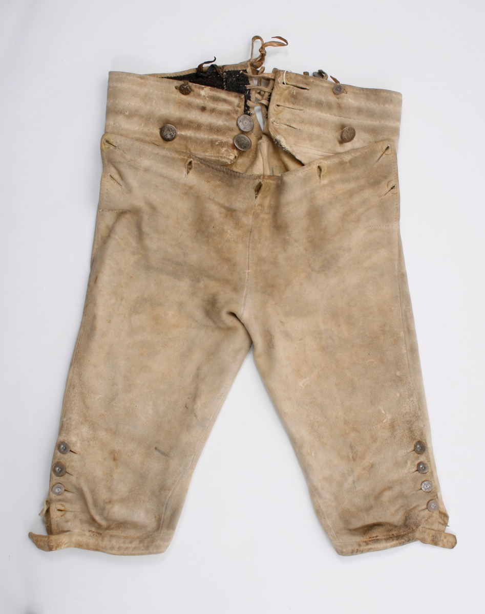 Håndsydd knebukse i lyst skinn. Lav midjeplassering. Linningen i midjen og lokket lukkes med ulike og graverte messingfargede knapper.  I høyre side ved linningen er det innsydd en lomme. I linningen MB er det splitt som lukkes med skinnsnor gjennom 3 x 2 hull og MF lukkes den med 3 knapper. Linningen er foret med ullstoff og har 4 rader prikkestikning gjennom for og skinn. Stor vidde over baken. Nede ved knærne er det splitter som lukkes med 4 knapper. Påsydd knelinning som trolig har hatt spenne for lukking. Alle knappene er festet med skinnsnor som igjen er festet på innsiden av plagget.Alle sammensyingskanter har tittekanter.