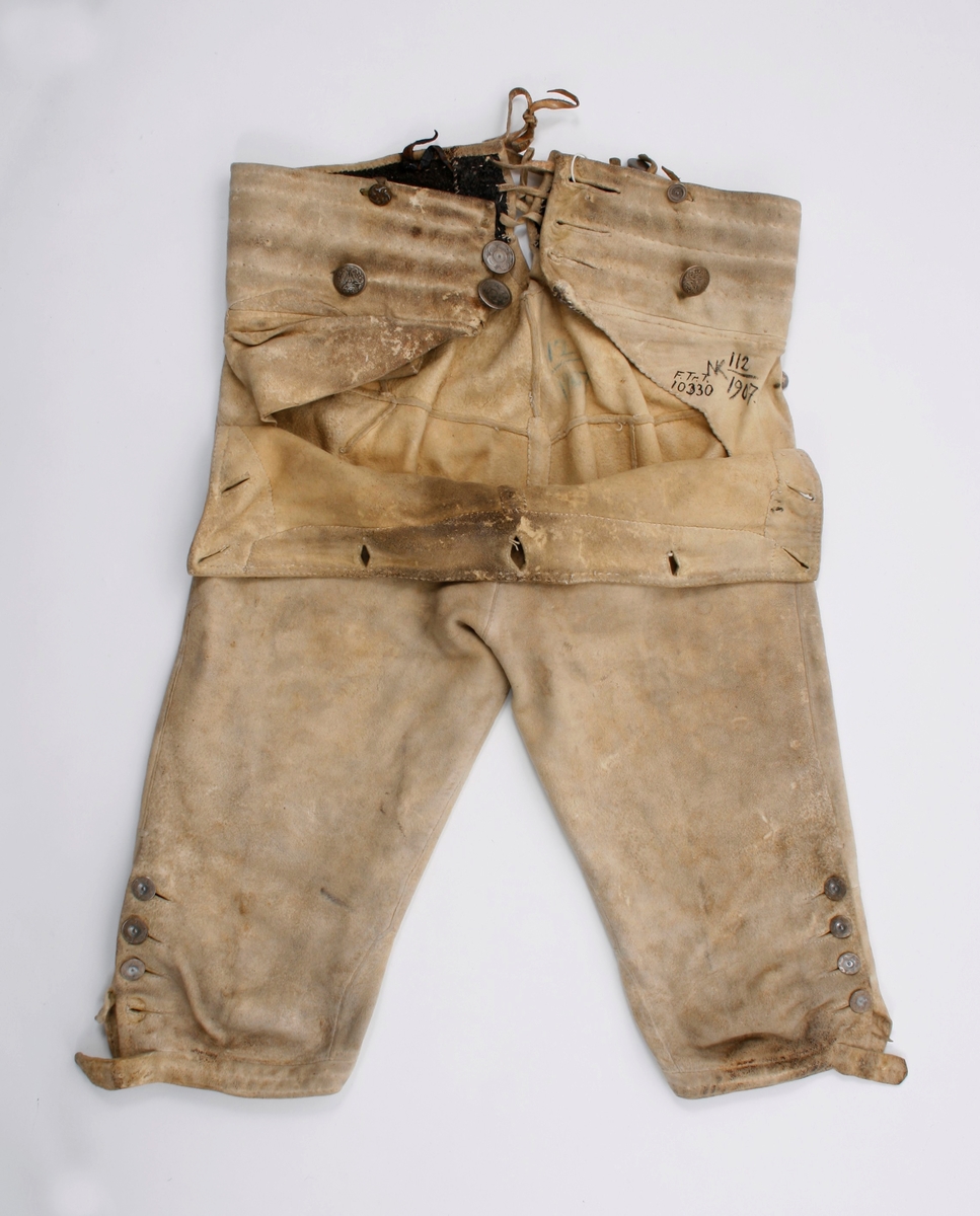 Håndsydd knebukse i lyst skinn. Lav midjeplassering. Linningen i midjen og lokket lukkes med ulike og graverte messingfargede knapper.  I høyre side ved linningen er det innsydd en lomme. I linningen MB er det splitt som lukkes med skinnsnor gjennom 3 x 2 hull og MF lukkes den med 3 knapper. Linningen er foret med ullstoff og har 4 rader prikkestikning gjennom for og skinn. Stor vidde over baken. Nede ved knærne er det splitter som lukkes med 4 knapper. Påsydd knelinning som trolig har hatt spenne for lukking. Alle knappene er festet med skinnsnor som igjen er festet på innsiden av plagget.Alle sammensyingskanter har tittekanter.