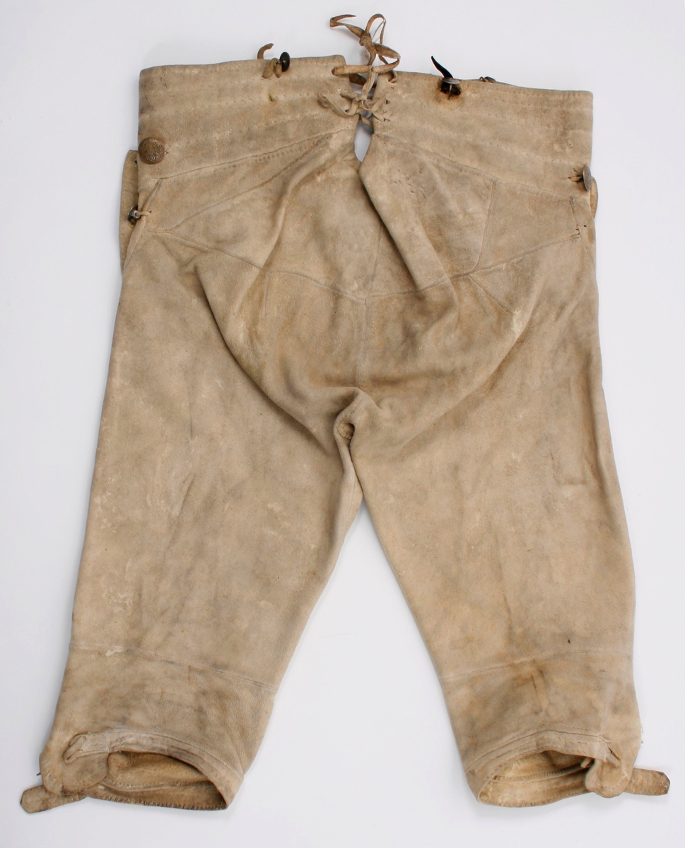 Håndsydd knebukse i lyst skinn. Lav midjeplassering. Linningen i midjen og lokket lukkes med ulike og graverte messingfargede knapper.  I høyre side ved linningen er det innsydd en lomme. I linningen MB er det splitt som lukkes med skinnsnor gjennom 3 x 2 hull og MF lukkes den med 3 knapper. Linningen er foret med ullstoff og har 4 rader prikkestikning gjennom for og skinn. Stor vidde over baken. Nede ved knærne er det splitter som lukkes med 4 knapper. Påsydd knelinning som trolig har hatt spenne for lukking. Alle knappene er festet med skinnsnor som igjen er festet på innsiden av plagget.Alle sammensyingskanter har tittekanter.