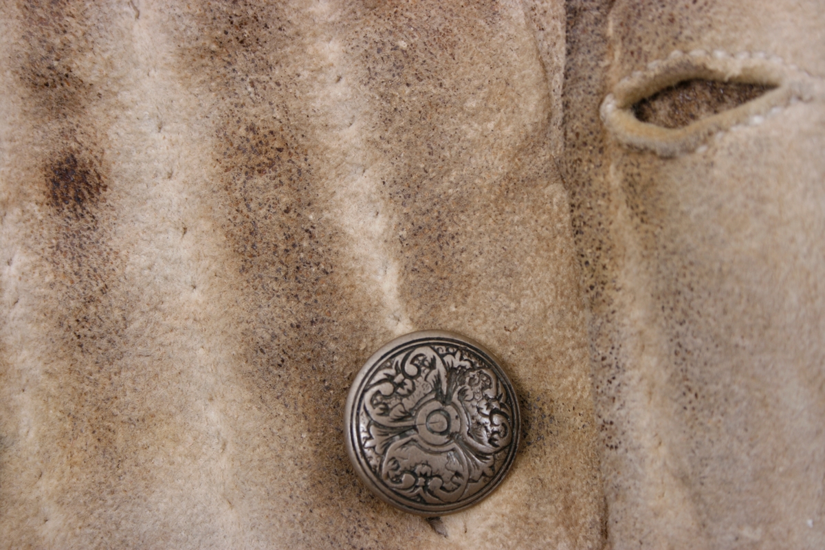 Håndsydd knebukse i lyst skinn. Lav midjeplassering. Linningen i midjen og lokket lukkes med ulike og graverte messingfargede knapper.  I høyre side ved linningen er det innsydd en lomme. I linningen MB er det splitt som lukkes med skinnsnor gjennom 3 x 2 hull og MF lukkes den med 3 knapper. Linningen er foret med ullstoff og har 4 rader prikkestikning gjennom for og skinn. Stor vidde over baken. Nede ved knærne er det splitter som lukkes med 4 knapper. Påsydd knelinning som trolig har hatt spenne for lukking. Alle knappene er festet med skinnsnor som igjen er festet på innsiden av plagget.Alle sammensyingskanter har tittekanter.