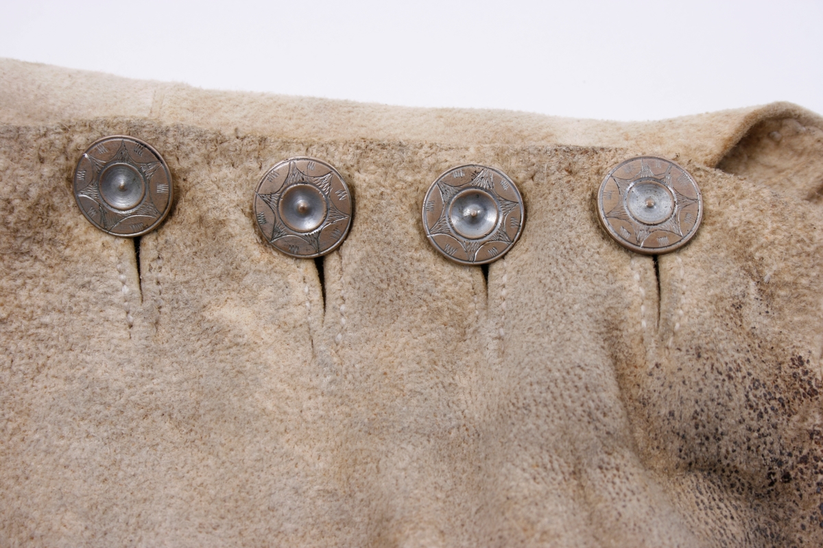 Håndsydd knebukse i lyst skinn. Lav midjeplassering. Linningen i midjen og lokket lukkes med ulike og graverte messingfargede knapper.  I høyre side ved linningen er det innsydd en lomme. I linningen MB er det splitt som lukkes med skinnsnor gjennom 3 x 2 hull og MF lukkes den med 3 knapper. Linningen er foret med ullstoff og har 4 rader prikkestikning gjennom for og skinn. Stor vidde over baken. Nede ved knærne er det splitter som lukkes med 4 knapper. Påsydd knelinning som trolig har hatt spenne for lukking. Alle knappene er festet med skinnsnor som igjen er festet på innsiden av plagget.Alle sammensyingskanter har tittekanter.