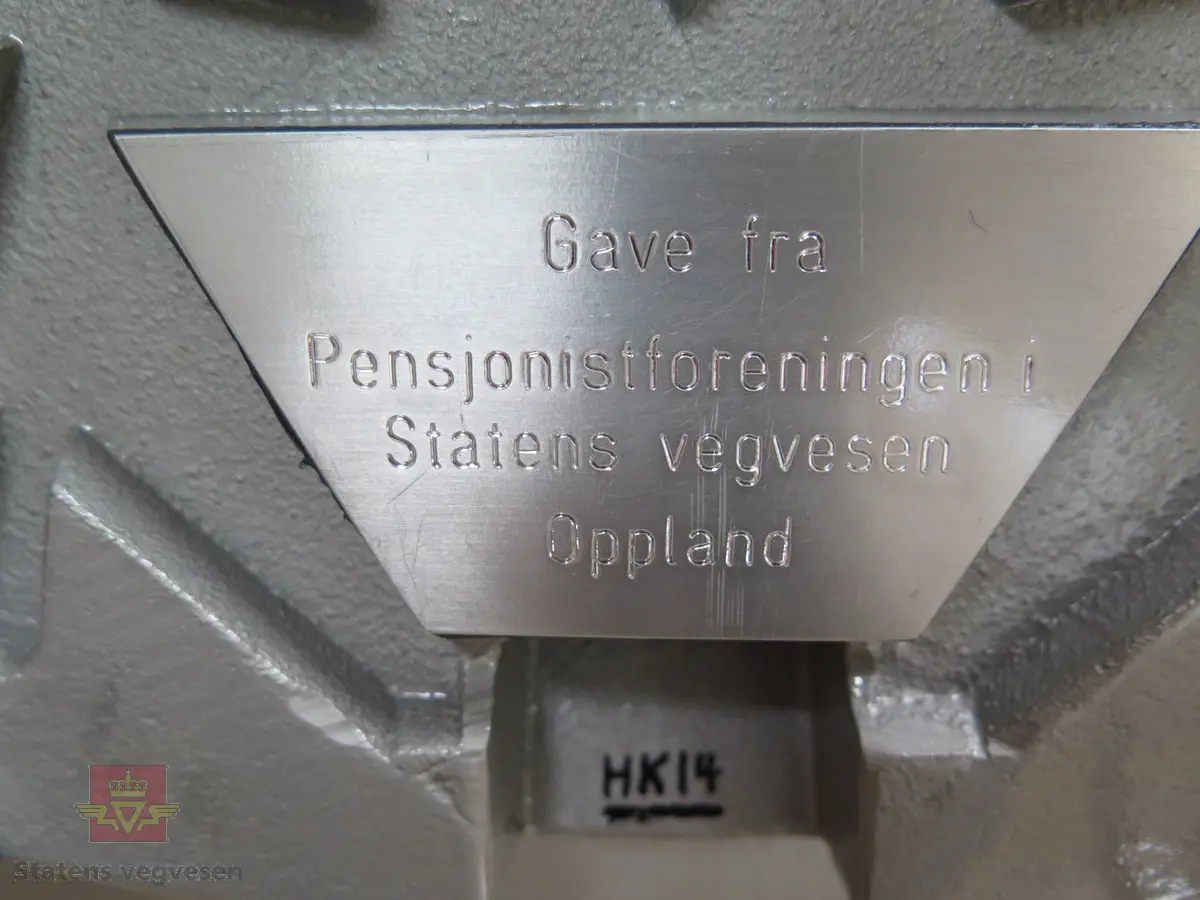 Kumlokk med vegvesenets logo. Lokket er malt i sølv, blått og svart. Logoen er malt rød og gul. Med lokket følger det er sølvfarget stativ av tre som lokket kan plasseres på. Lokket har merking fra produsent og giver.