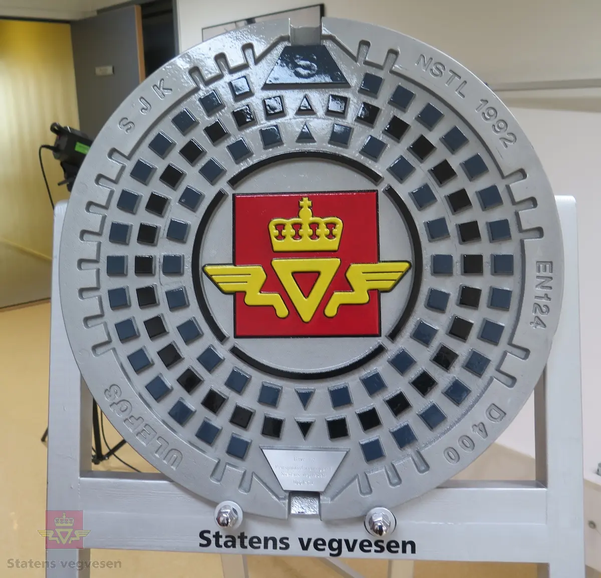 Kumlokk med vegvesenets logo. Lokket er malt i sølv, blått og svart. Logoen er malt rød og gul. Med lokket følger det er sølvfarget stativ av tre som lokket kan plasseres på. Lokket har merking fra produsent og giver.