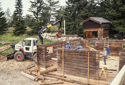 Tømmermaskin løfter en laftestokk mens fire blåkledde menn jobber med å lafte opp et gammelt laftehus.