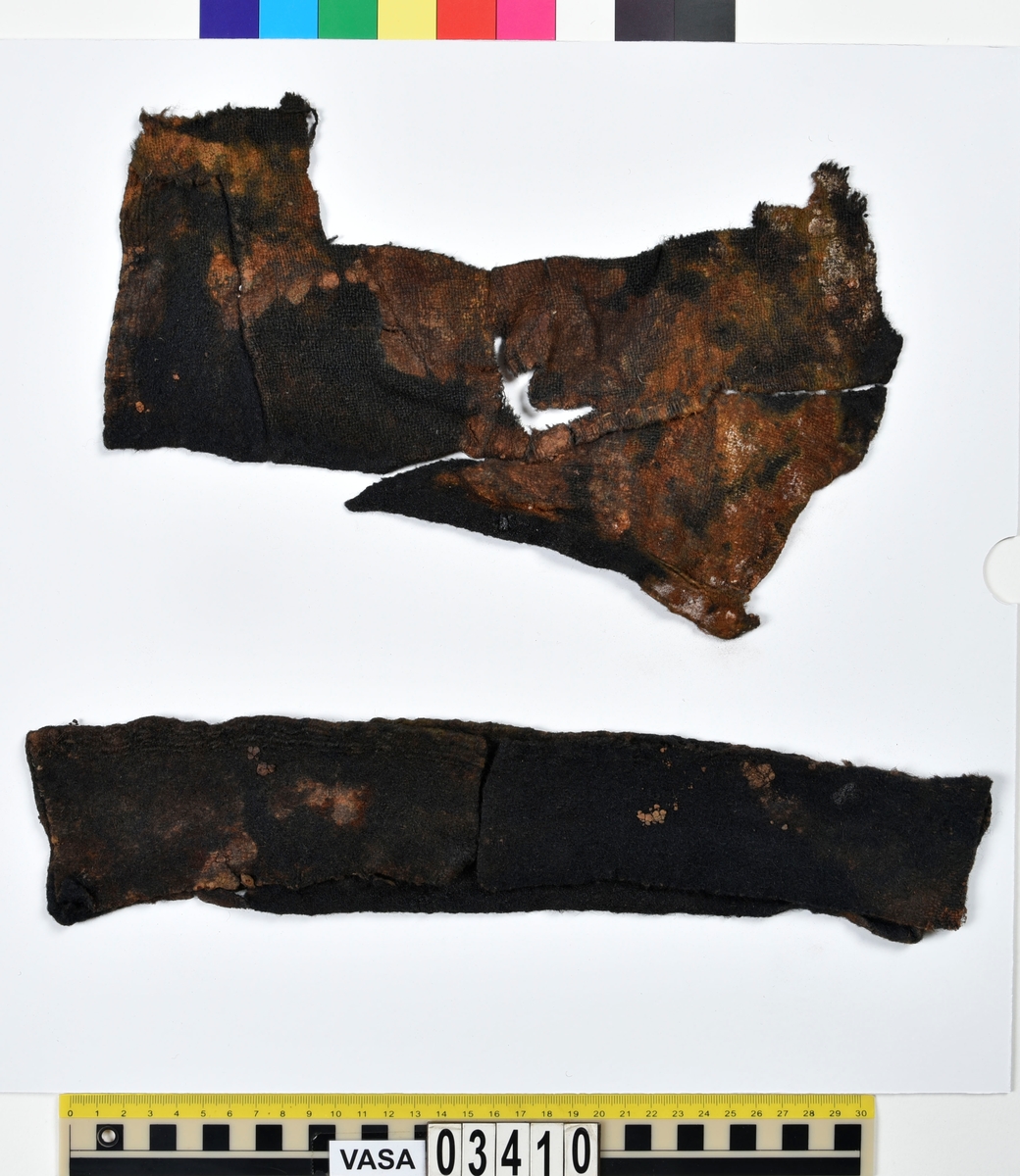 Textil.
3 fragment.
Ett fragment består av tre tygbitar hopsydda med en ljus tråd (lin?) till ett stycke. Ett annat fragment består av två tygbitar hopsydda med en ljus tråd (lin?) till ett stycke. Det tredje fragmentet är löst.
Tyget är vävt av ullgarn i tuskaft och är svartbrunt. Ser ut att ha varit valkat. En del av bitarna har stadkanter. Fragmenten har svarta blanka fläckar av tjära eller dylikt.