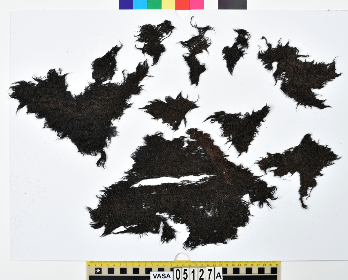 Textil.
Fyndnummer 05127 är uppdelat i a-e.
Fnr 05127a består av 9 fragment av ull vävda i 2/2-kypert. 
Fnr 05127b består av 8 fragment av ull vävda i tuskaft. Tyget är mer valkat på ena sidan än den andra. Två av fragmenten har delar av originalkanter, med spår av sömmar, kvar.
Fnr 05127c består av 4 fragment av ull vävda i tuskaft. Alla fragmenten har delar av originalkanter, med spår av sömmar, kvar.
Fnr 05127d består av ett fragment av ull vävt i tuskaft. Fragmentet en originalkant, med spår av söm, kvar.
Fnr 05127e består av 3 fragment av ull vävda i gles tuskaft.