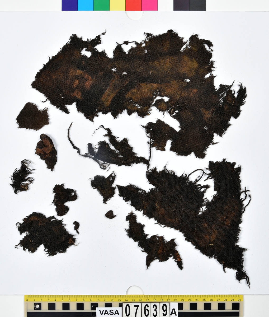 Textilfragment. 53 fragment fördelade på fyndnummer 07639a-e.
Fnr 07639a består av 17 fragment av ull vävda i tuskaft.
Fnr 07639b består av 8 fragment av hattfilt.
Fnr 07639c består av 17 fragment av ull vävda i kypert.
Fnr 07639d består av 1 fragment av ull vävt i kypert.
Fnr 07639e har 10 fragment av ull vävda i tuskaft varav ett fragment har en stadkant.

Textildokumentation 2017:
07639b
9 fragment i hårt filtad filt. Filten är tjock och kompakt, typ 1:1 har även en klippt eller skuren del. Sidan ett (synlig på bilden 1) är mycket mörkare i ytan, nästan svart, medan sida två går mer åt brunt. Båda sidorna har synliga märken efter avtryck, På sida två syns dessa bäst. En del hår i filten är finare än andra och dessa blänker mer. Möjligen är materialet bäver.
07639d:
Ett tygfragment i kompakt, tät och grov kypert, typ 3:1. Fragmentet har kraftigt upprepade kanter runtom. Tjocka fransar har bildats på den längsta sidan, dessa består av trådsystem 1. Trådsystem 1 är mycket hårt Z-tvinnat, nästan så att det lägger sig i öglor. Tyget är glansigt och på sida ett ser det något ripsartat ut, då trådsystem 1 nästan inte syns, utan bara randningen.  Sida två av tyget har nötningar och glesare kypertränder.