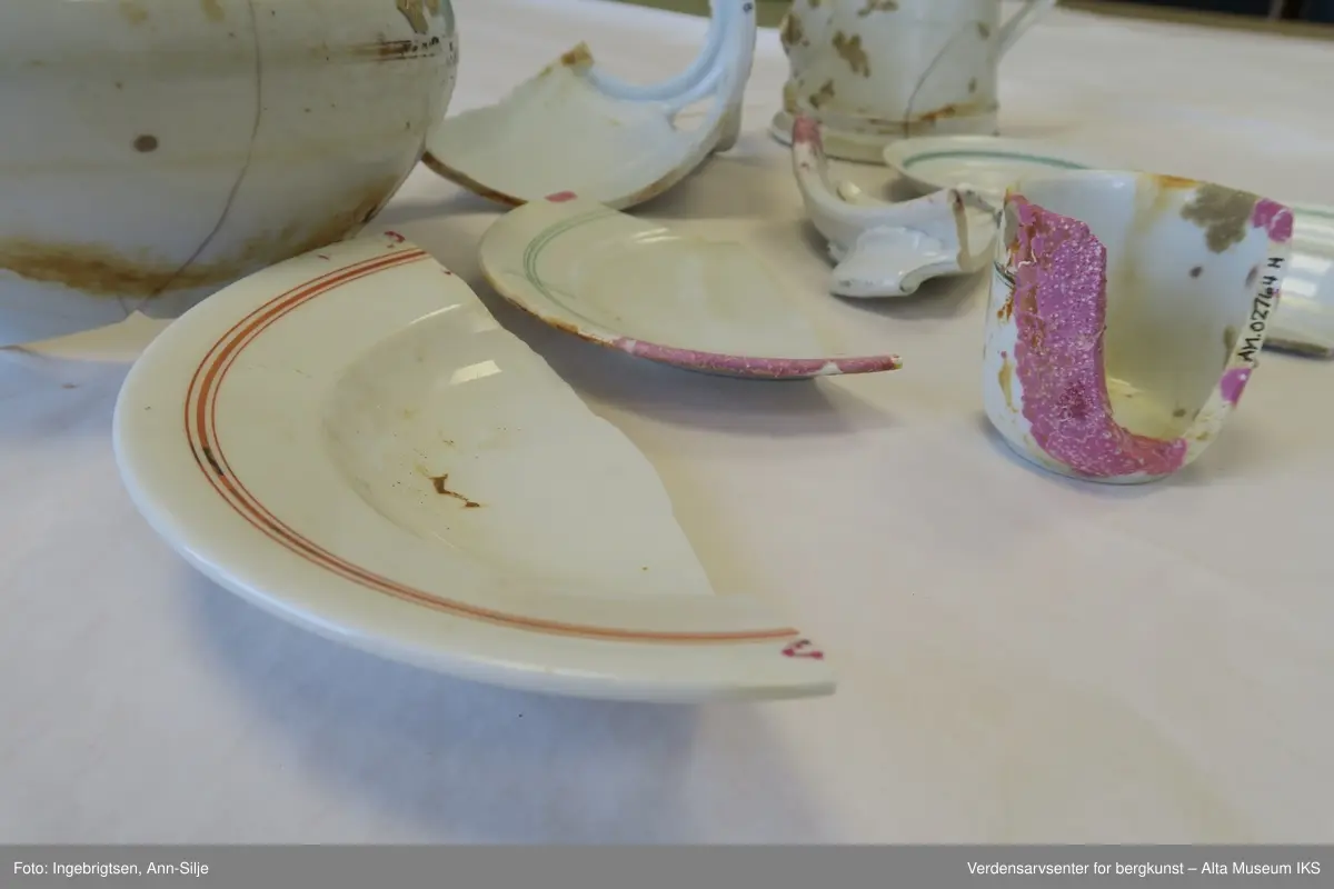 Bruddstykkene er av A) Terrin, B og C) Kanne, D) Dyptallerken, E) Flattallerken, F) Serveringsfat?, G og J) Tefat, H) Kaffekopp, I) Asjett. De er alle sammen hvite og noen av de har grønne eller røde striper rundt kantene. På undersiden av gjenstandene er det stempel med produsentnavn og hakekors med ørn over.