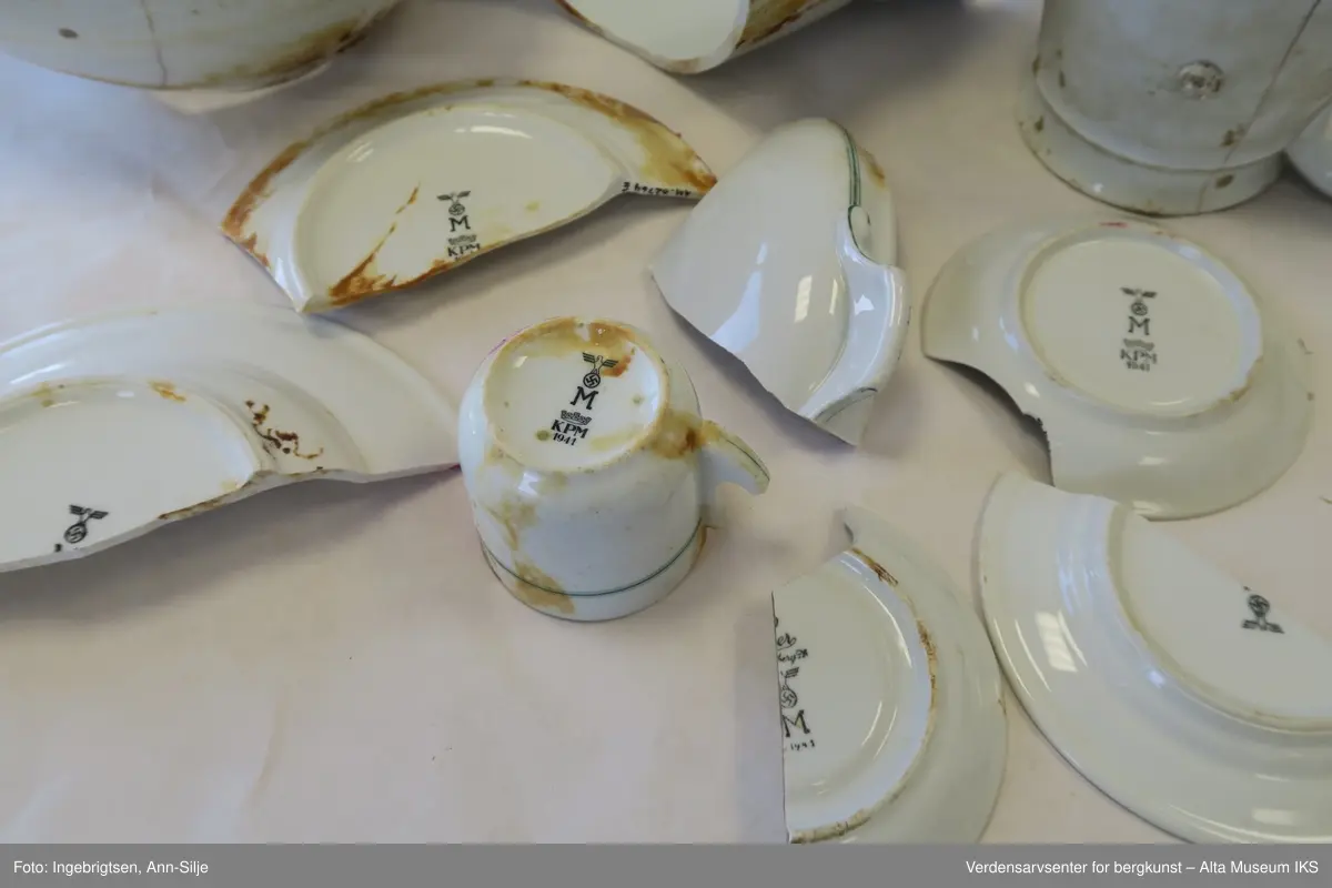 Bruddstykkene er av A) Terrin, B og C) Kanne, D) Dyptallerken, E) Flattallerken, F) Serveringsfat?, G og J) Tefat, H) Kaffekopp, I) Asjett. De er alle sammen hvite og noen av de har grønne eller røde striper rundt kantene. På undersiden av gjenstandene er det stempel med produsentnavn og hakekors med ørn over.